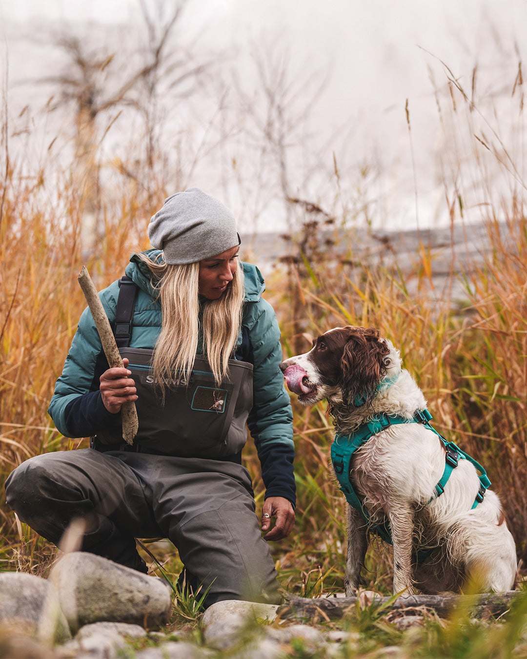 Paula Shearer playing with a stick with her dog Willow on the side of the Bow River. Her dog is wearing the Saker Muse harness in Skeena Teal