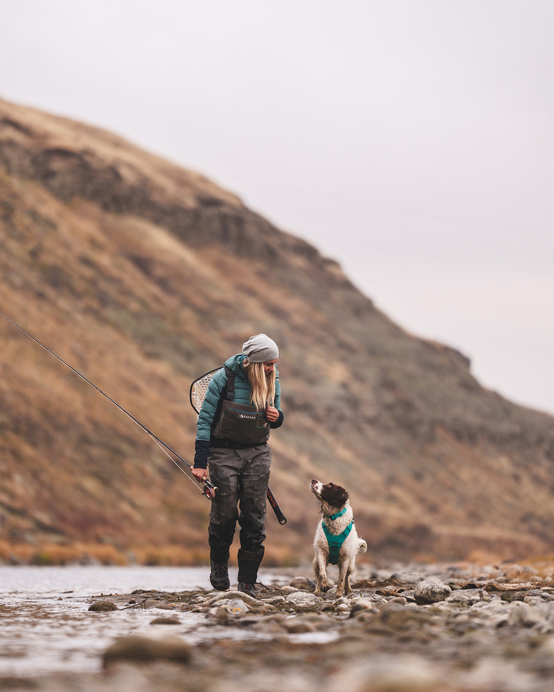 Paula Shearer with her dog Willow walking on the side of the Bow River. Her dog is wearing the Saker Muse harness in Skeena Teal